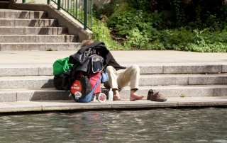 Photographie d'une personne en situation d'itinérance avec de nombreux bagages aux abords d'une étendue d'eau